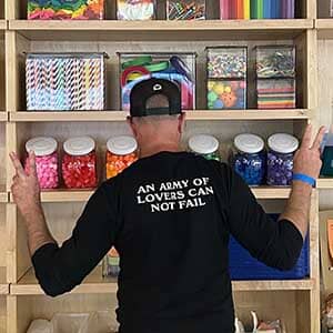 Person wearing a black t-shirt with text standing in front of shelves filled with colorful containers.