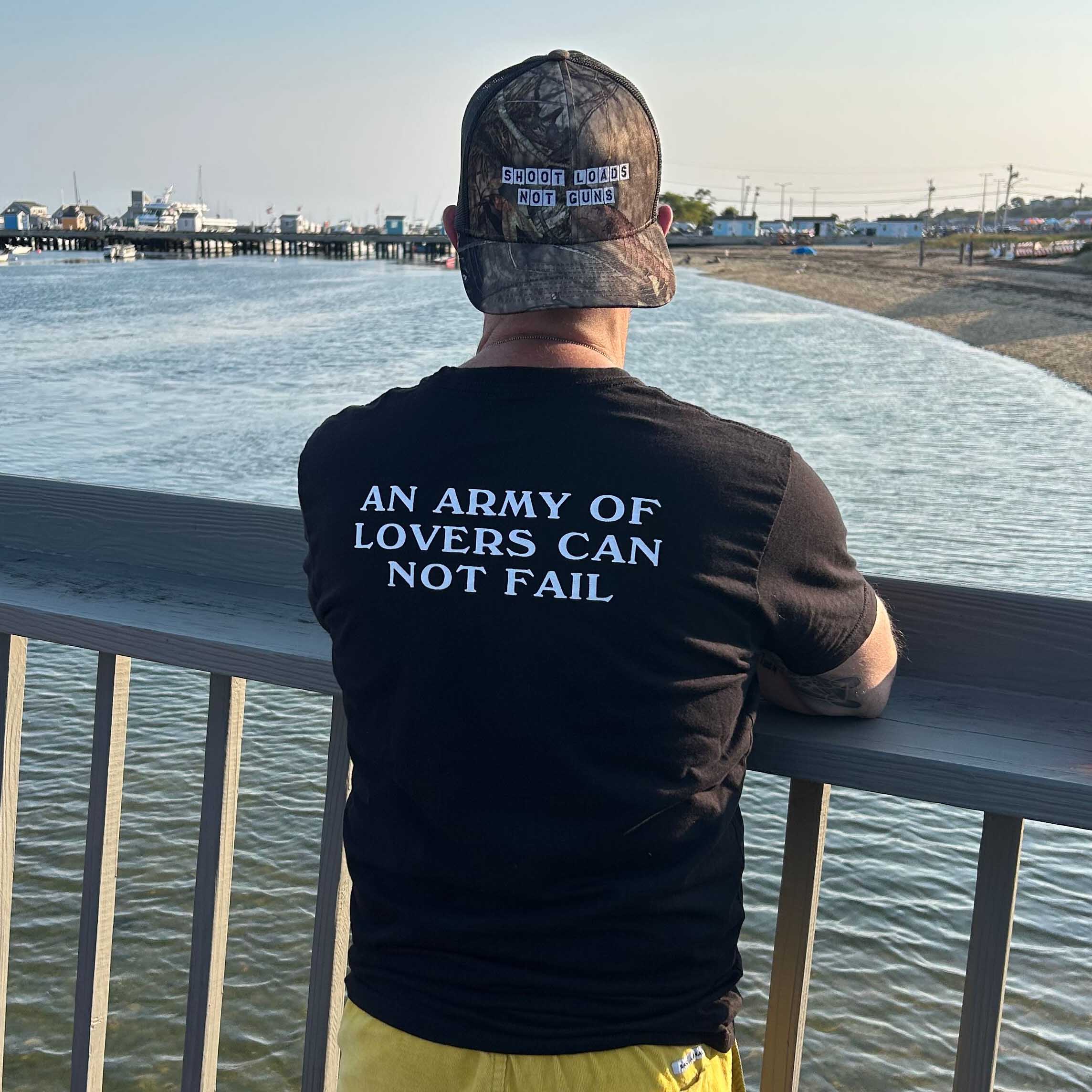 Person wearing a black t-shirt with text and a cap, standing by a waterfront.