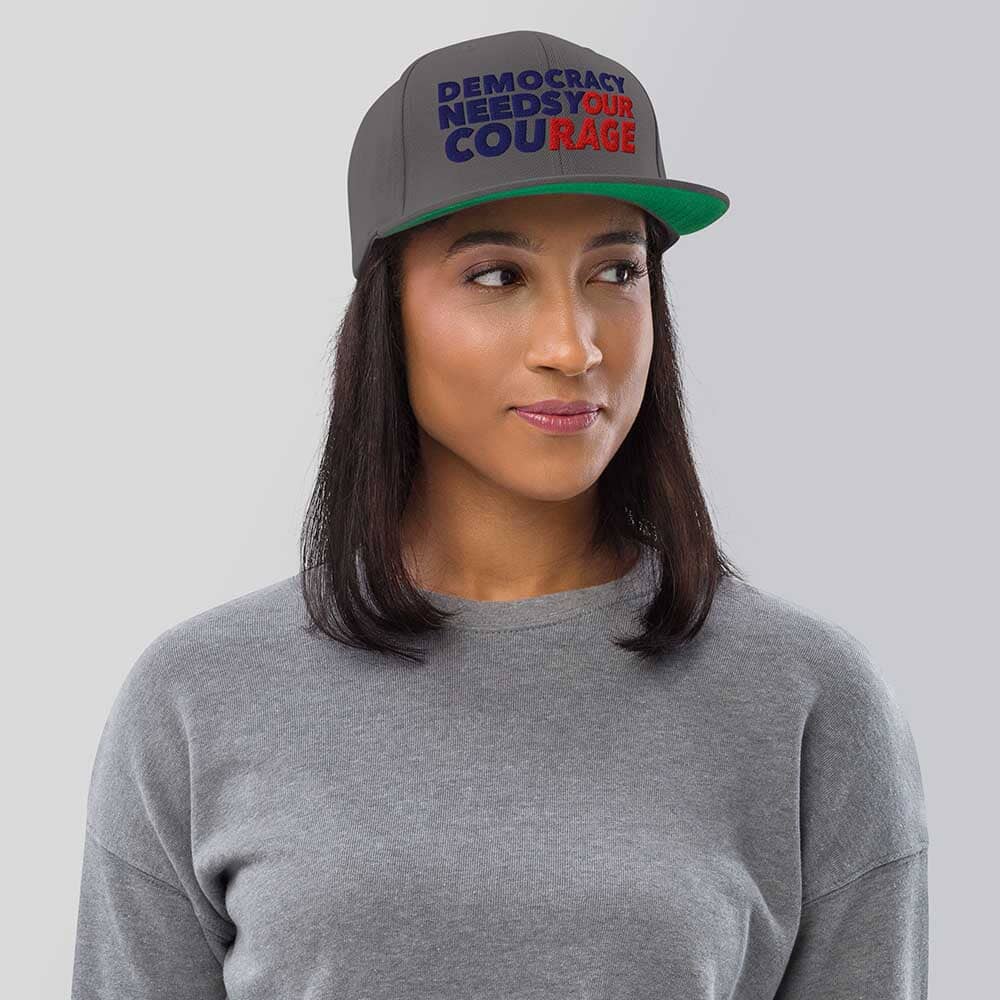 Person wearing a cap with 'Democracy Needs Your Courage' text on a plain background