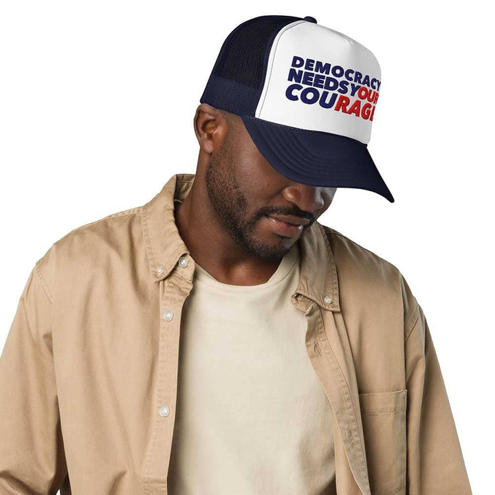 Man wearing a navy and white cap with DEMOCRACY NEEDS YOUR COURAGE, beige jacket, and beige shirt on a white background