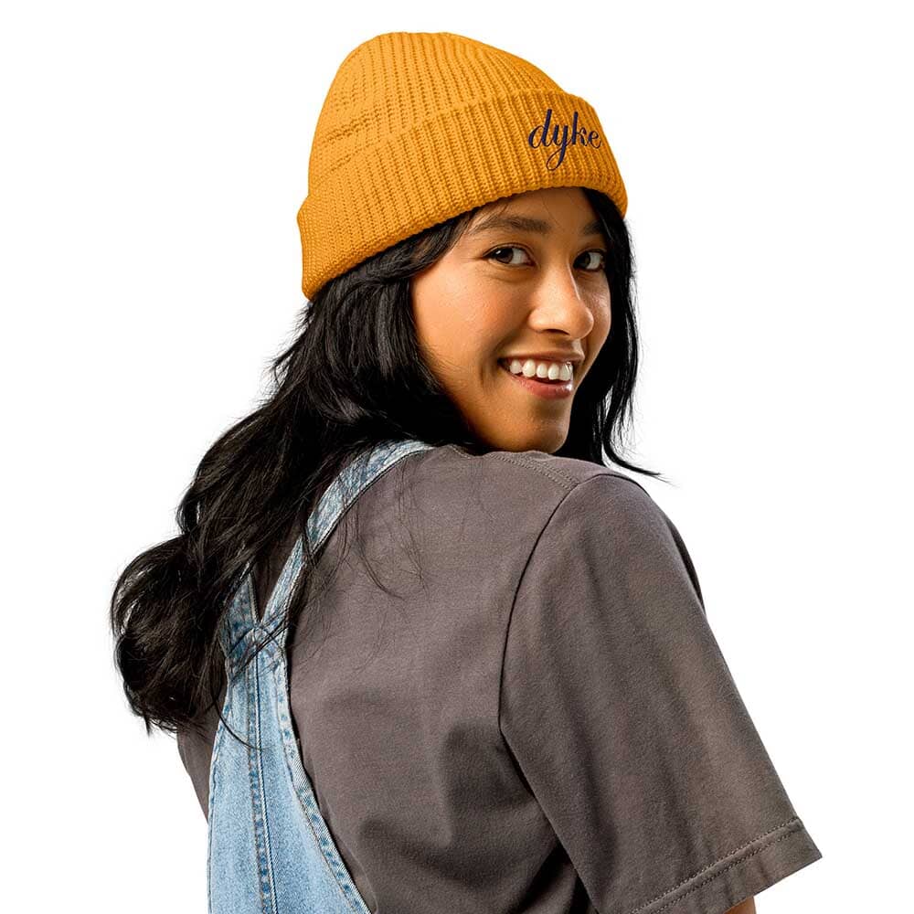 Person wearing a mustard yellow beanie with 'dyke' text on a white background
