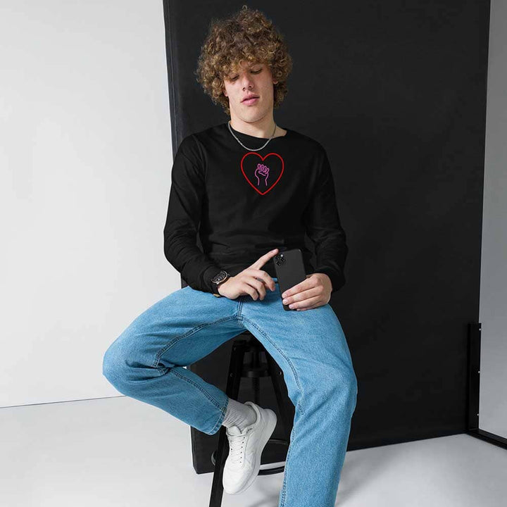 Person wearing a black sweatshirt with a red heart design and pink solidarity fist, sitting on a chair against a black backdrop.