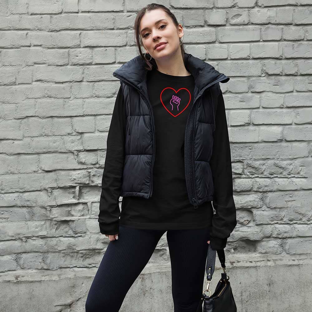 Woman wearing a black puffer vest over a black shirt with a heart and pink solidarity fist design, standing against a brick wall.