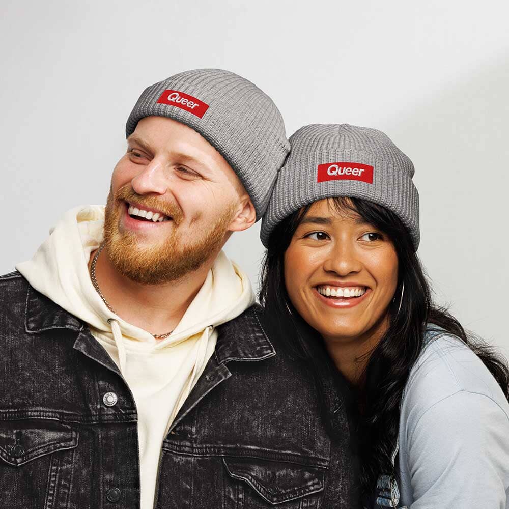 Two people wearing gray 'Queer' beanie hats against a plain background