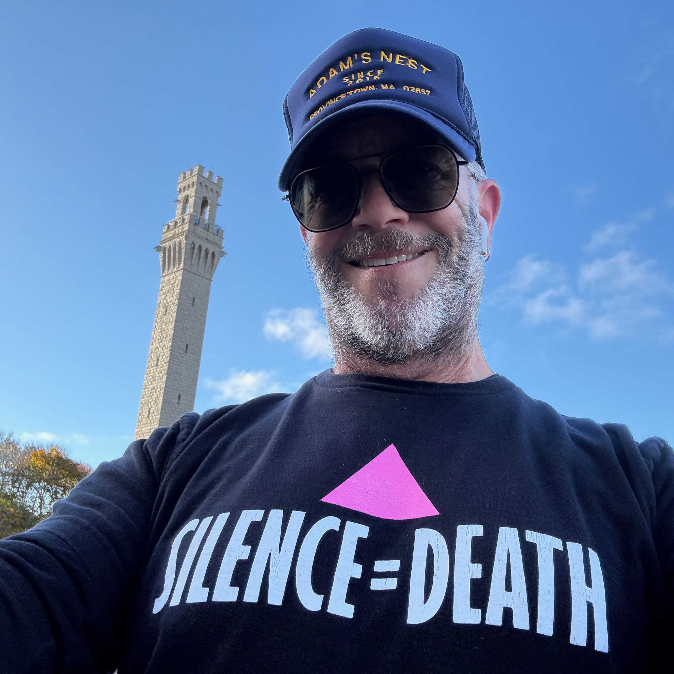Adam Singer wearing a cap and  silence equals death t-shirt and sunglasses with provincetown pilgrim monument in background