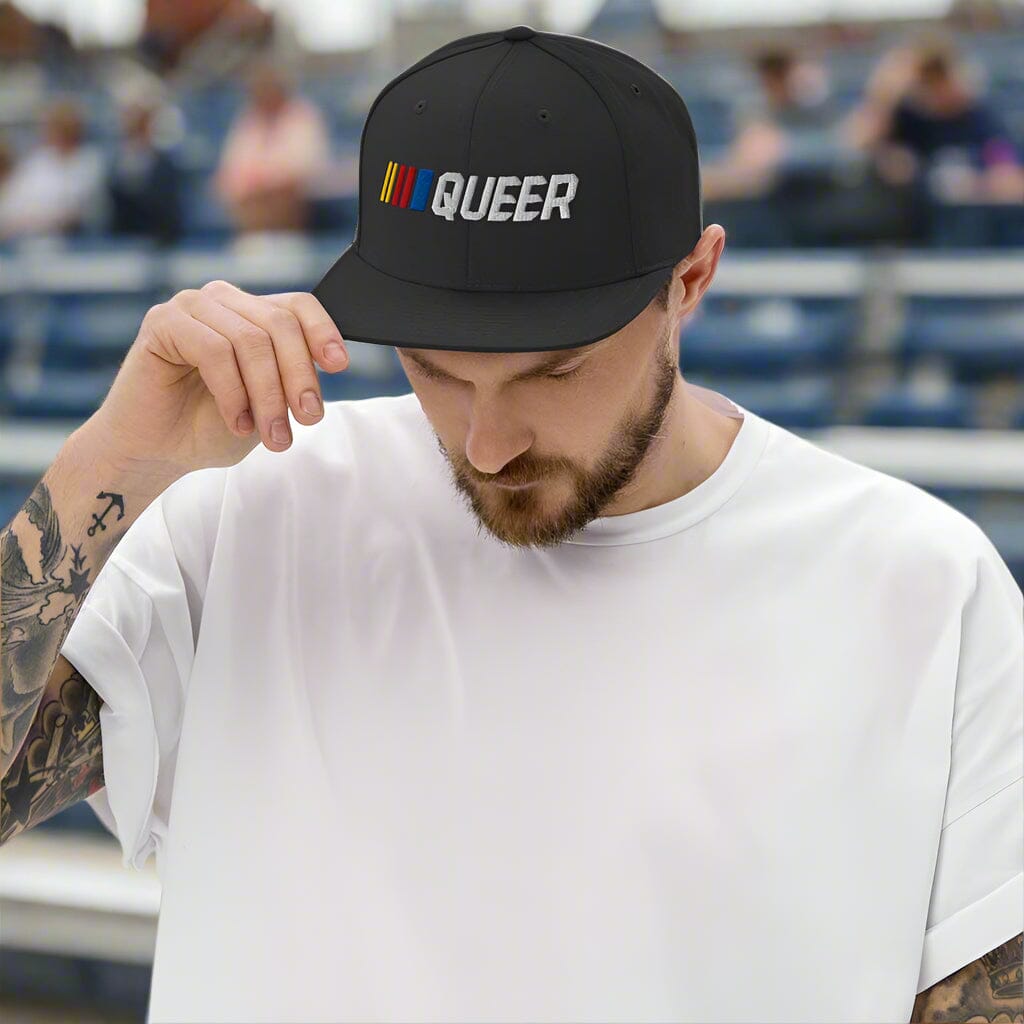 Man wearing a black cap with 'QUEER' text and stripes in stands