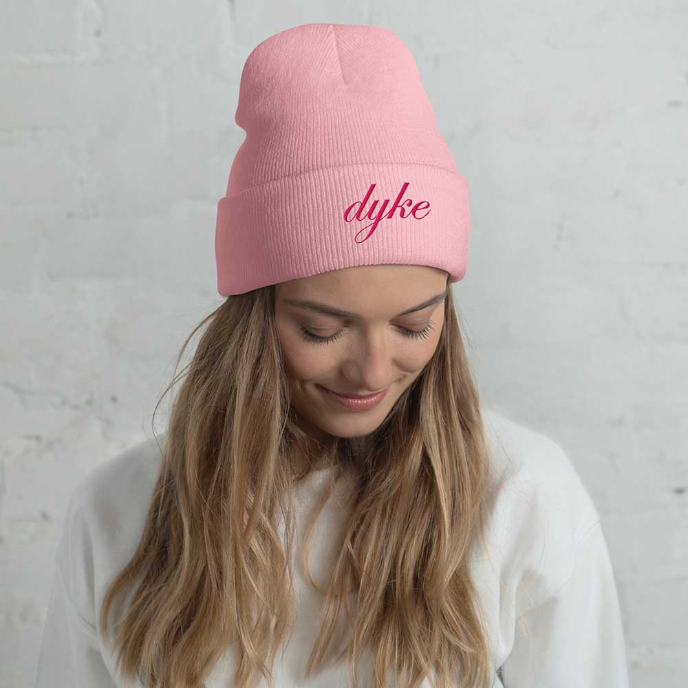 person wearing a pink beanie with 'dyke' text against a gray background