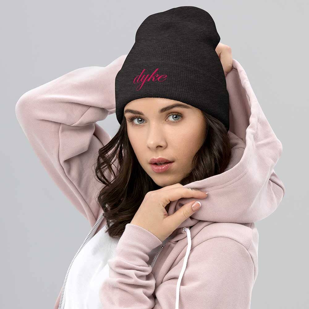 Woman wearing a charcoal grey beanie with 'dyke' written on it and a pink hoodie.