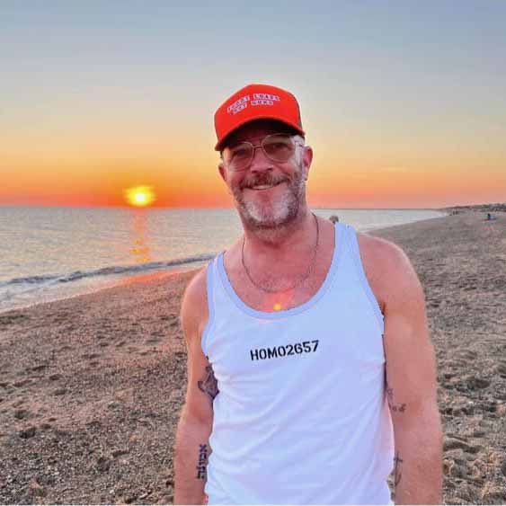 adam singer wearing a blaze orange cap reading shoot loads not guns and white tank top with 'HOMO2657' on a beach at sunset.