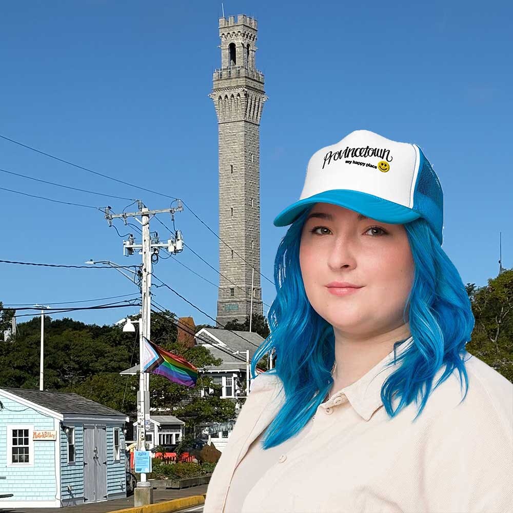 Person with blue hair wearing a 'Provincetown' cap in front of a tall tower and clear blue sky.